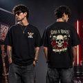 Two men modeling black Guns N' Roses t-shirts in a dark, industrial setting. The man on the left shows the front of the shirt with a small yellow and white graphic, while the man on the right shows the back of the shirt featuring a large graphic of a skull and roses with the Guns N' Roses logo.