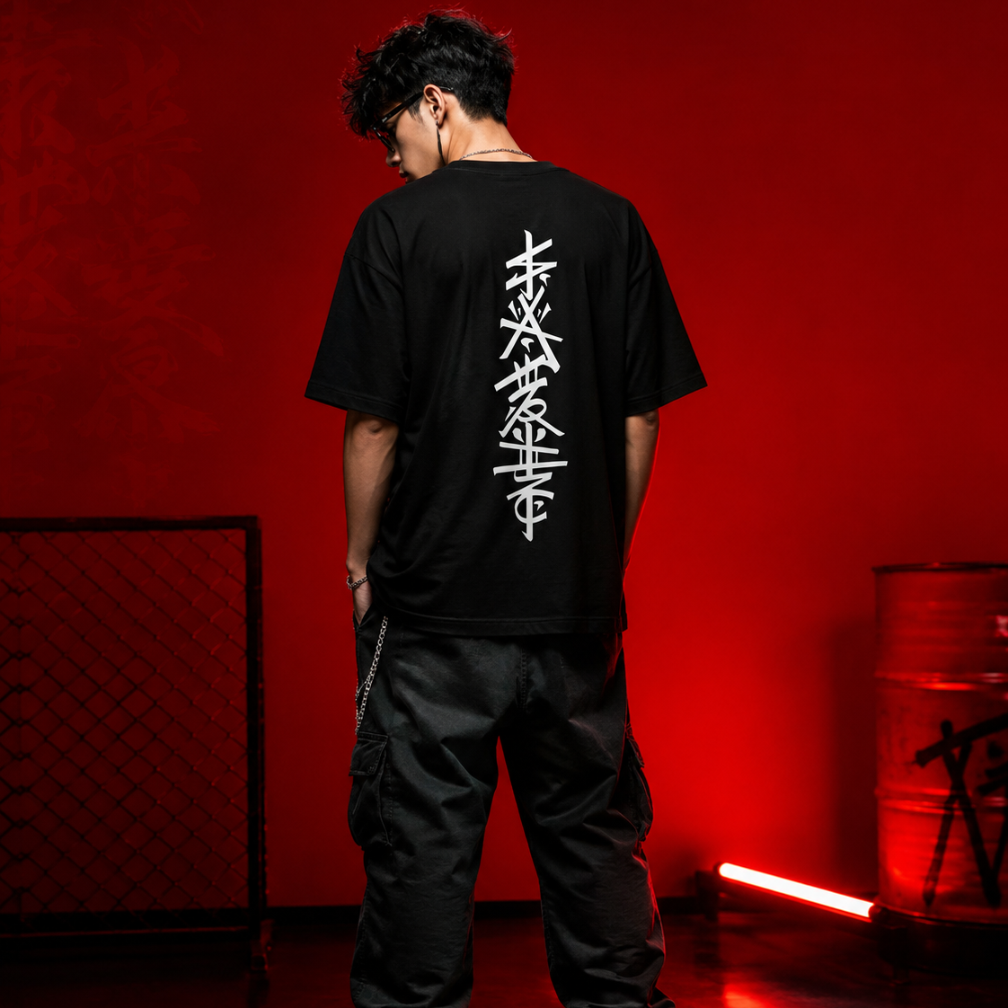 A man stands with his back to the camera wearing a black t-shirt with a large white vertical graphic print on the back, styled with black cargo pants and black-and-white sneakers in a dark studio setting with red lighting.