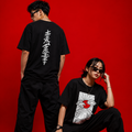Two models posing in black graphic t-shirts and black pants against a solid red studio background. One model stands with their back to the camera, showing a vertical white graphic print, while the other crouches in front wearing a graphic t-shirt with a red heart and white geometric design.